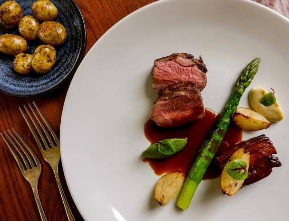 A fine dining plate with sliced lamb, asparagus, roast potatoes, vegetables, and a red wine reduction, served beside a dish of seasoned baby potatoes.
