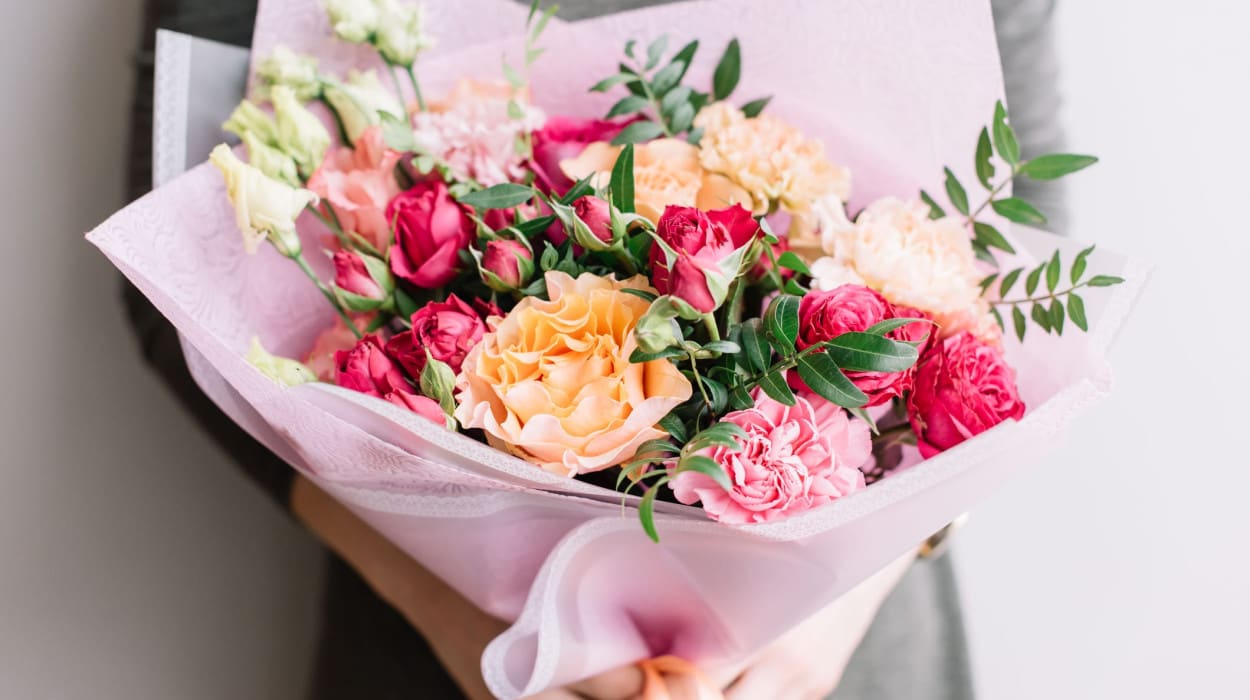 A person in a gray outfit holds a bouquet of pink, red, and peach roses and carnations, wrapped in soft pink paper with greenery accents.