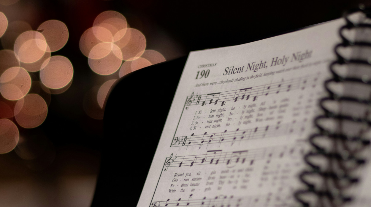 Open hymn book showing “Silent Night, Holy Night” sheet music on a piano stand, with warm Christmas lights softly blurred in the background.