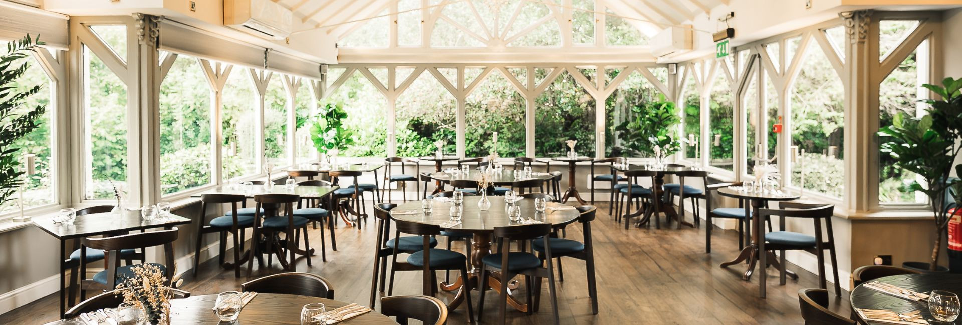 Bright conservatory-style restaurant with wooden floors, dark tables, and blue chairs surrounded by large windows overlooking lush garden greenery.